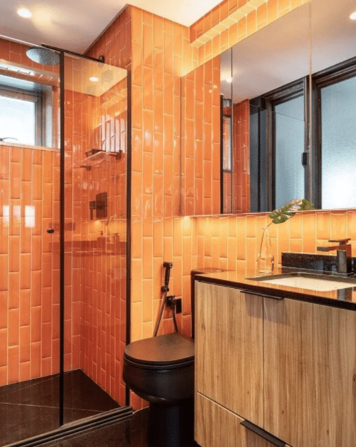 Banheiro com revestimento laranja subway tiles na parede, piso e louças pretas e armário amadeirado 