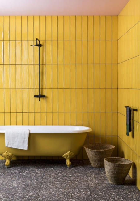 Banheiro com azulejo subway tile amarelo na parede, banheira imersa amarela, piso porcelanato reproduzindo mármore cinza e cestos de palha no chão