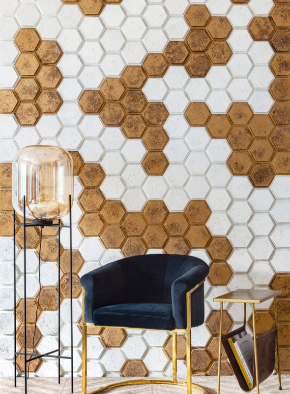 Ambiente com poltrona de estofado azul, mesinha de canto dourada, luminária de chão, de fundo, parede revestida de peças hexagonais brancas e douradas.