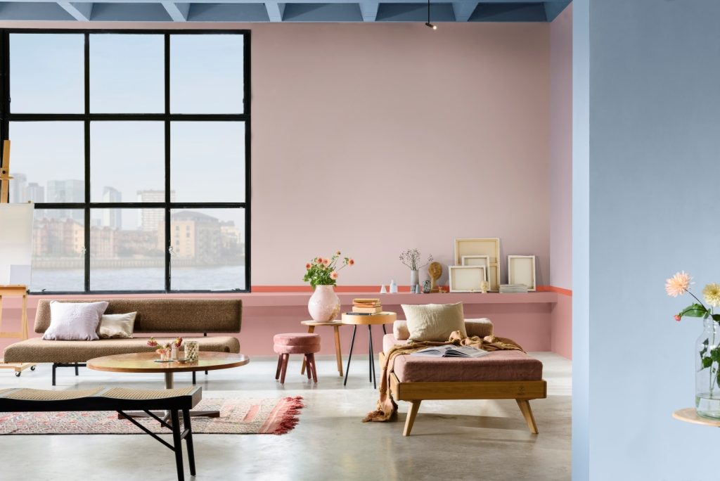 Sala com parede principal rosa claro, paredes laterais azul clara, piso em cimento queimado, sofás e banquetas marrons, mesinhas de canto coloridas com vasos.