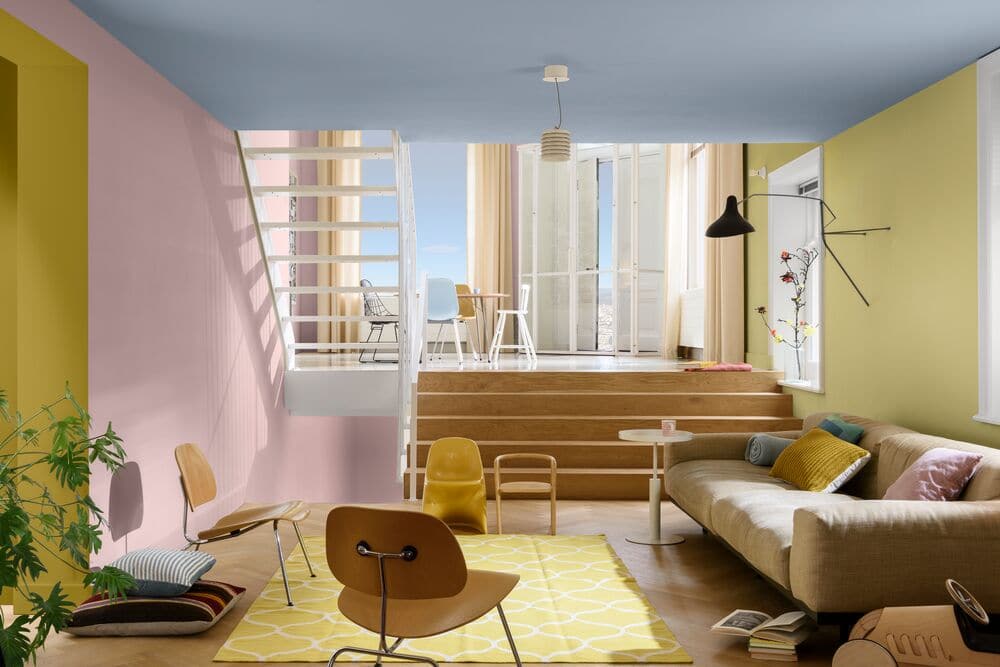 Sala com predes coloridas rosa e amarelo, teto azul claro, piso bege, tapete amarelo, sofá e cadeiras bege.