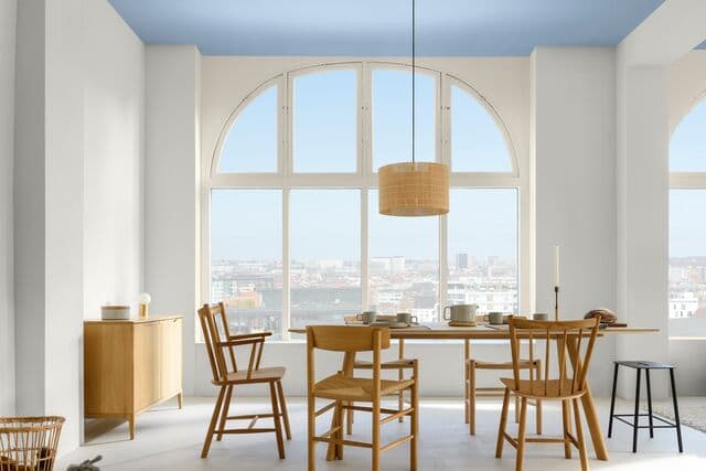 Ambiente com paredes e janelas brancas, teto azul claro, mesa e cadeiras de madeira, luminária pendente de palha, aparador de madeira no canto da parede.