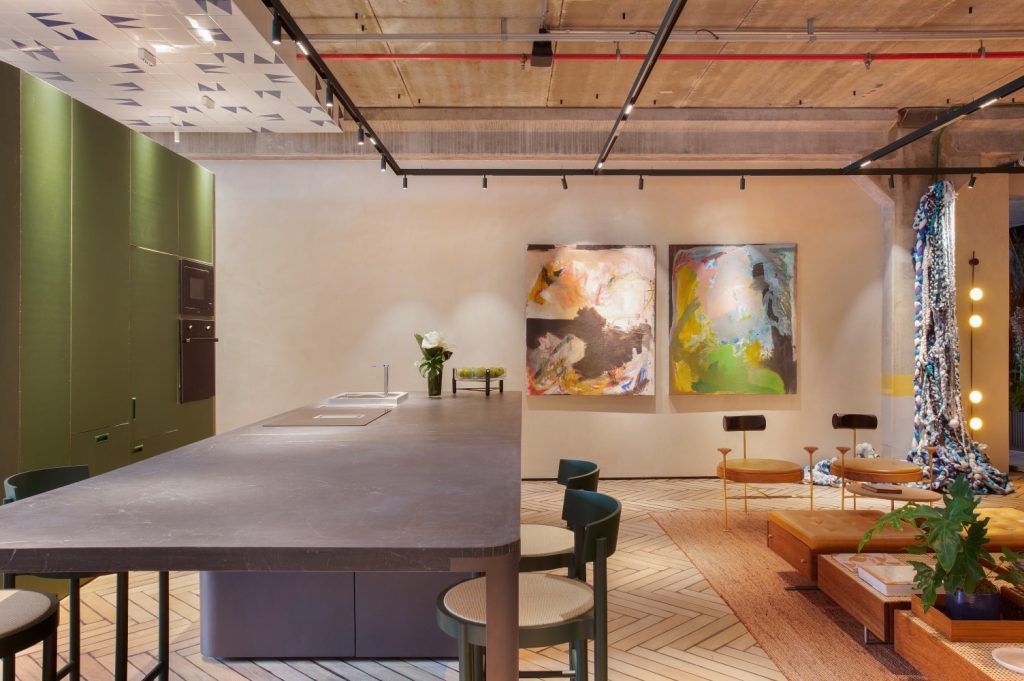 Cozinha com piso de madeira, mesa retangular de jantar cinza, armários verde integrado a sala de estar com mesinhas de madeira, poltrona de madeira com estofado