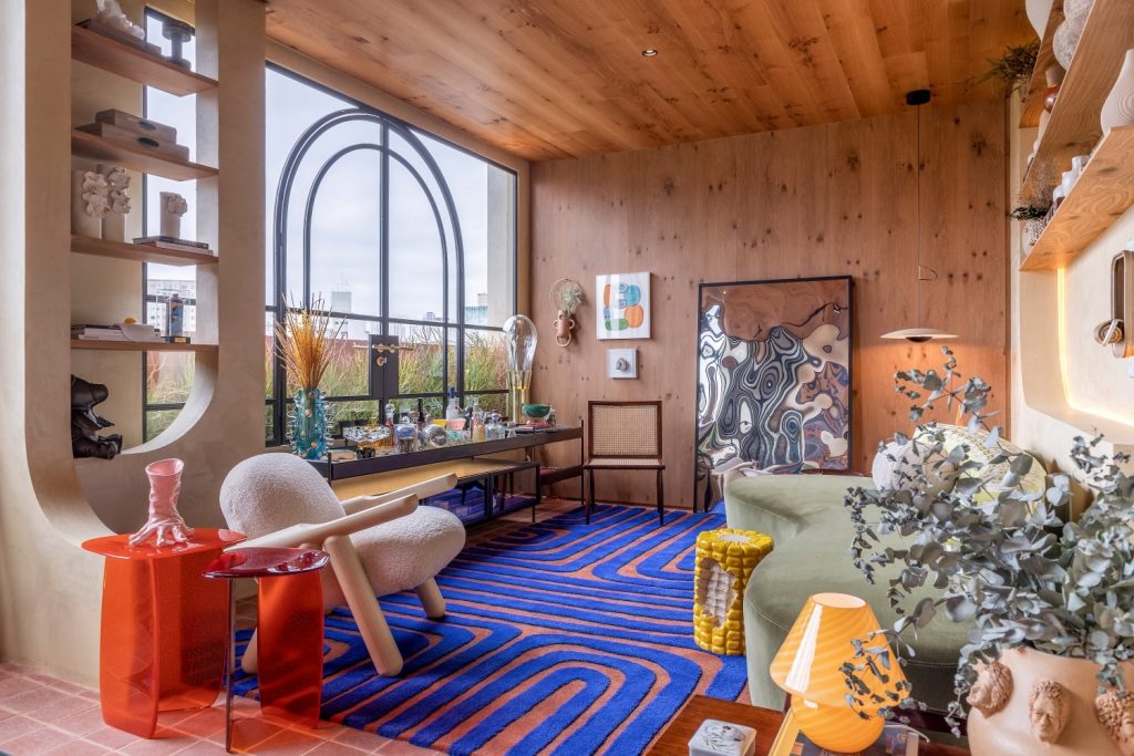 Living Galeria: Ambiente com revestimento de madeira na parede e teto, janela grande com detalhes na estrutura de ferro, tapete azul e marrom, piso de cerâmica, sofá verde, poltrona branca, mesinhas de centro laranjas e prateleiras com muitos objetos 