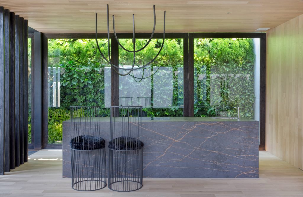 Vidros Vivix: Bancada revestida com material marmorizado cinza escuro, duas cadeiras de ferro com estofado azul escuro, piso, parede e teto com revestimento de madeira e portas grandes envidraçadas