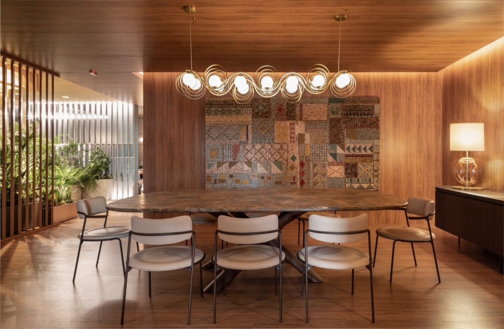 Sala de jantar com piso, paredes e teto revestido de madeira, mesa de jantar oval com oito cadeiras com estofado nude, luminária pendente comprida sob a mesa e painel colorido na parede central