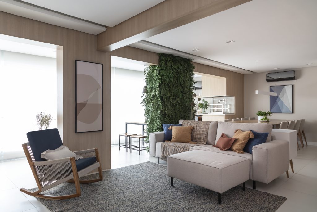 Sala de estar com integrada a sala de jantar com sofá branco, poltrona de madeira com estofado azul, nuances de madeira clara, piso porcelanato branco e jardim vertical