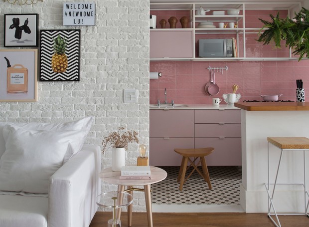 Vista da sala de estar com parede de tijolinhos branca, piso de madeira, sofá branco, de fundo cozinha com azulejo rosa, armários rosa claro, bancada de madeira com banquetas de madeira
