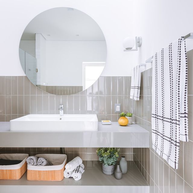 Banheiro com azulejo em meia parede na cor neutra vertical, e outra metade pintada de branco, espelho redondo, bancada pia em cor neutra com cuba branca