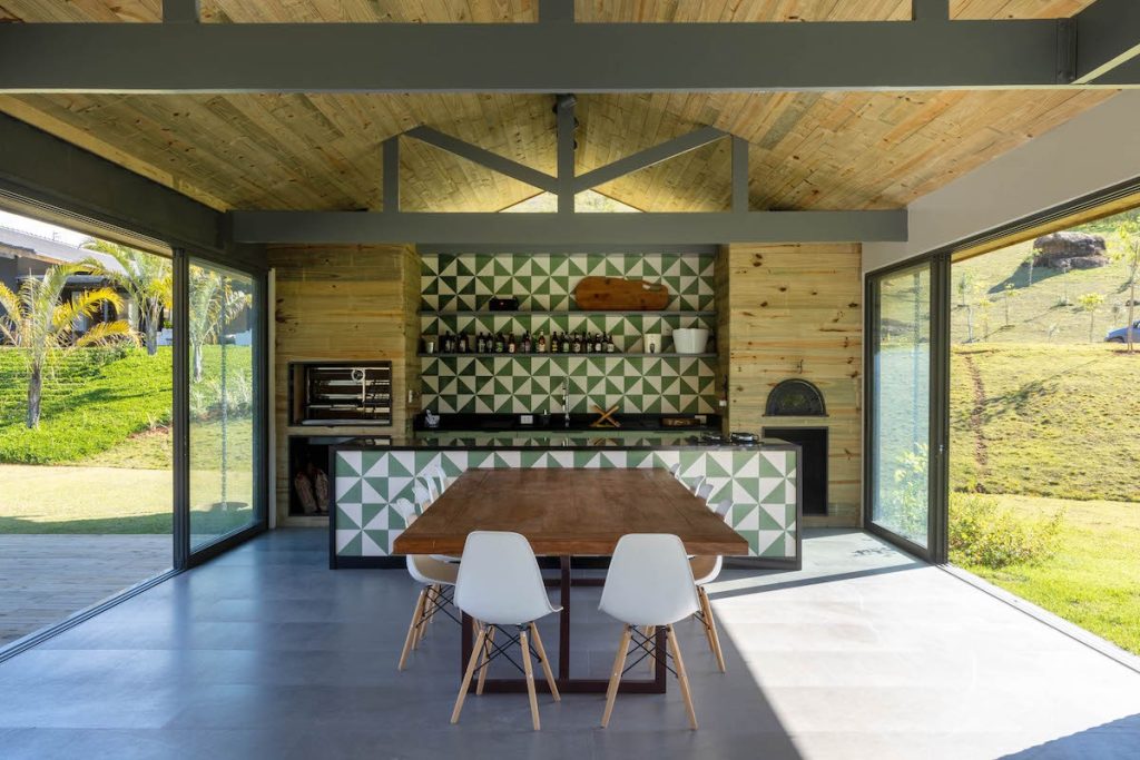 Area gourmet com teto e parede revestido de madeira, frontão da pia e bancada revestidos de ladrilhos hidráulicos verde e branco, mesa de madeira retangular com cadeiras brancas
