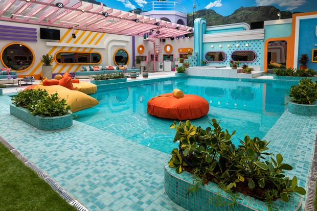 Área externa do BBB 22 com piscina. sofás, cadeiras, puffs e decoração em laranja, azul e rosa