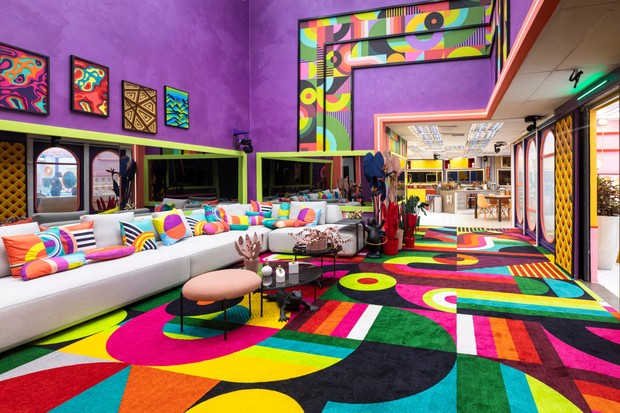 Sala do BBB 22 com tapetes coloridos, sofá branco com almofadas coloridas, parede roxa com quadros e elementos coloridos