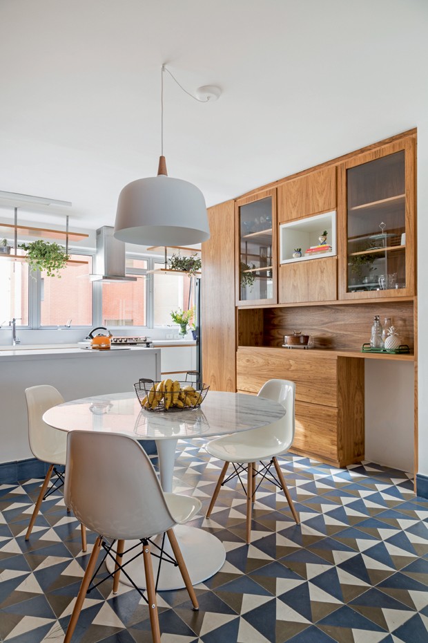 Cozinha com piso de ladrilho hidráulico azul e cinza, mesa redonda branca com três cadeiras brancas, luminária pendente branca sob a mesa, armário amadeirado, bancadas e paredes brancas