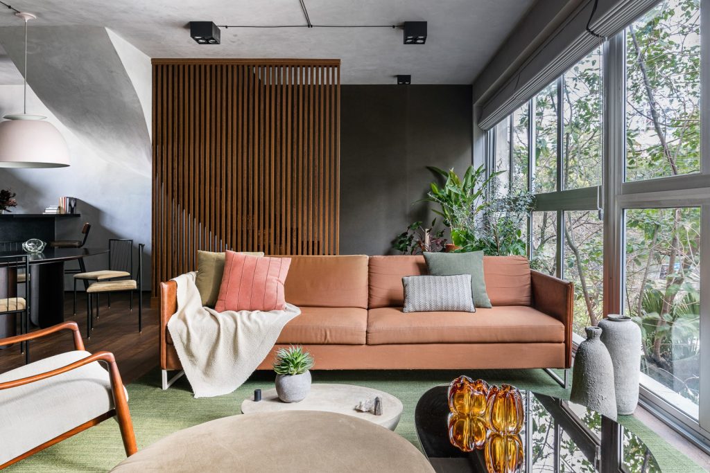 Sala com sofá laranja, tapete verde, divisória de madeira vazada, janela de vidro ponta a ponta na parede, piso de madeira, poltrona e mesinha de centro redonda