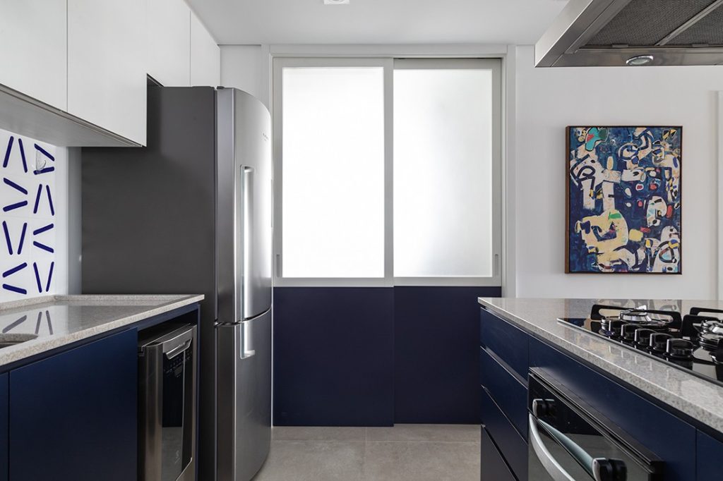Cozinha com marcenaria azul marinho, paredes brancas, parede da pia revestida com azulejo azul e branco, eletrodoméstico de inox e piso em tom cinza