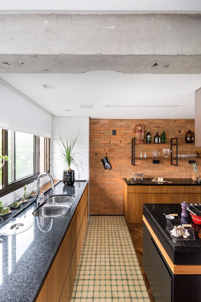 Cozinha com parede de tijolinhos, teto com viga de concreto aparente, armários amadeirado, bancada preta e piso de ladrilho hidráulico laranja