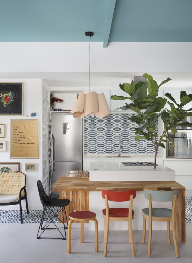 cozinha com piso em tom cinza, ladrilho hidraulico no frontão da pia azul e branco, mesa de madeira com 4 cadeiras coloridas, luminária pendente e plantas decorativas