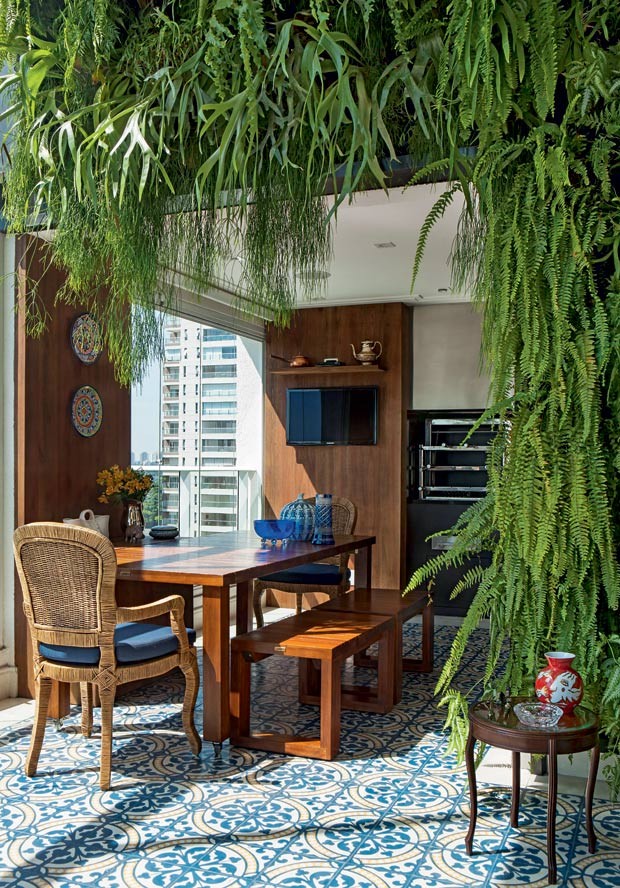Área gourmet com piso de ladrilho hidráulico azul e branco, mesa de madeira com bancos e cadeiras e plantas fazendo meio arco no ambiente.