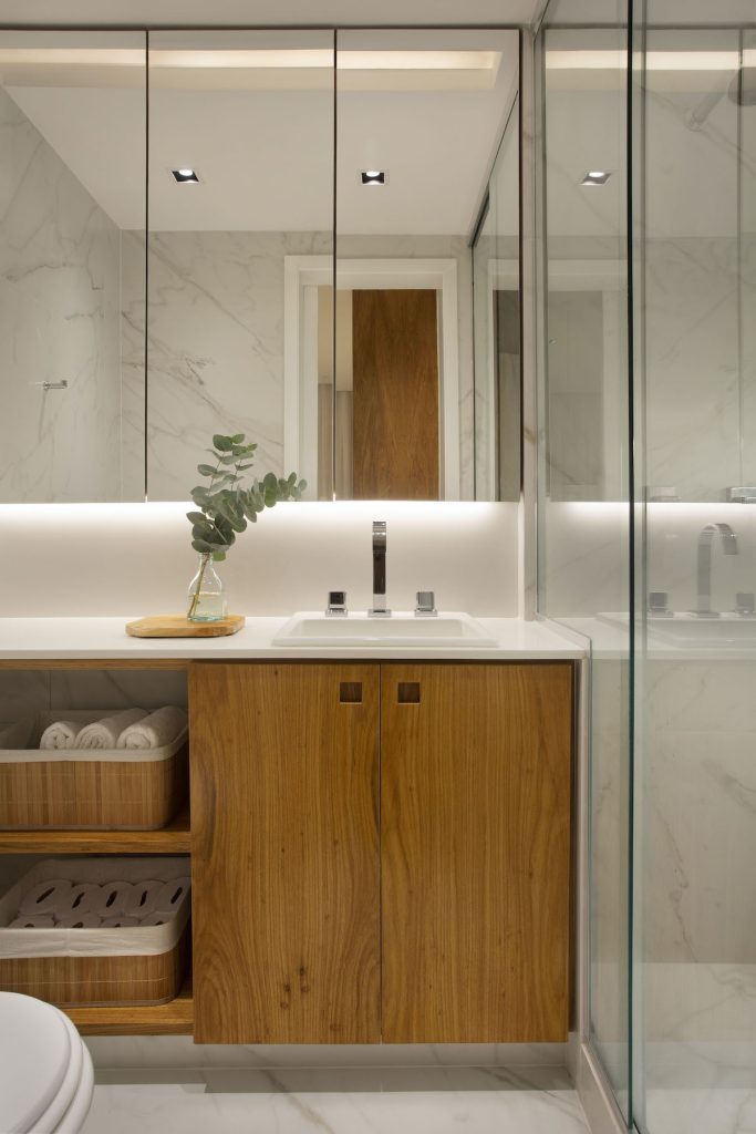 Banheiro revestido com porcelanatos imitando mármore, armário de madeira, bancada branca, box envidraçado