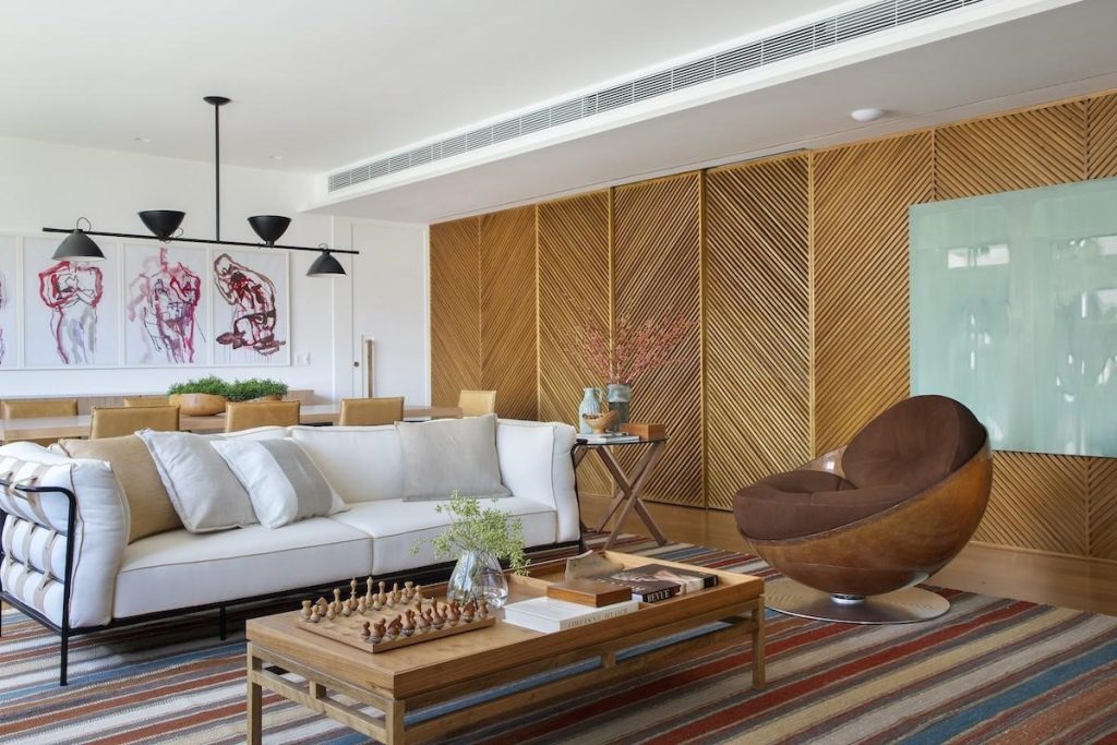 Sala de estar integrada com sala de jantar, com painéis ripados, sofá branco, poltrona marrom, tapete listrado,, mesinha de centro de madeira