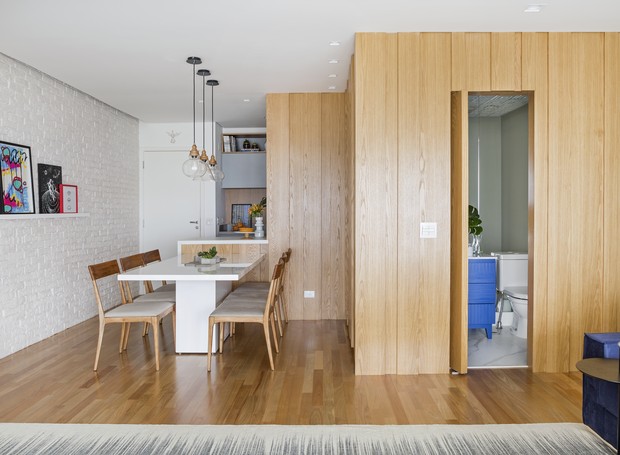 Sala de jantar e demais áreas de apartamento com tacos de madeira no piso, paredes com revestimento de madeira, mesa de jantar branca com luminárias pendentes