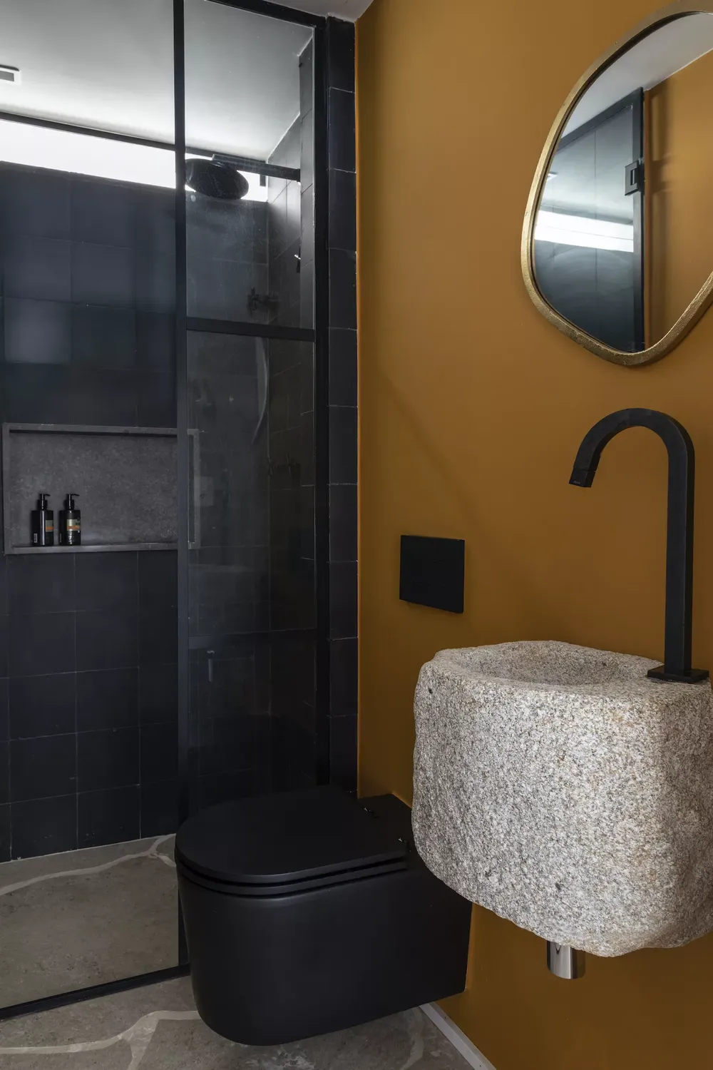 Banheiro com revestimento do box preto, louças pretas, pia de pedra. uma parede principa amarela com espelho organico